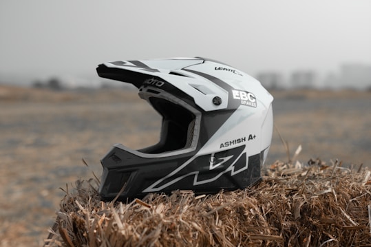 A black and white motocross helmet is resting on a bale of hay in an outdoor setting with a blurred background. The helmet has various brand logos and text decals, featuring sharp geometric designs.