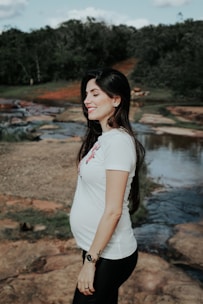 A pregnant woman stands outdoors by a rocky stream, smiling and wearing a white t-shirt and black pants. The background features green trees and a clear sky.