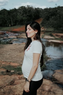 A pregnant woman stands outdoors by a rocky stream, smiling and wearing a white t-shirt and black pants. The background features green trees and a clear sky.