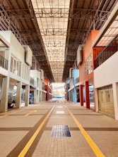 An indoor walkway is flanked by buildings with various colored facades, including shades of blue, orange, and white. The overhead ceiling is composed of a metal lattice structure, allowing natural light to filter through. The ground is tiled with a geometric pattern, highlighted by yellow lines, leading towards a distant open area.