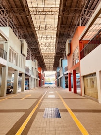 An indoor walkway is flanked by buildings with various colored facades, including shades of blue, orange, and white. The overhead ceiling is composed of a metal lattice structure, allowing natural light to filter through. The ground is tiled with a geometric pattern, highlighted by yellow lines, leading towards a distant open area.
