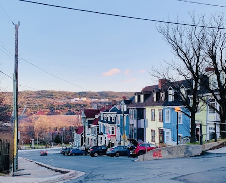 A quiet street lined with colorful historic row houses, featuring shades of blue, yellow, pink, and red. Leafless trees stand along the sidewalk, and a distant hillside creates a scenic backdrop. Several cars are parked along the street, and the word 'LOVE' is painted on a concrete barrier. Overhead wires stretch across the sky, which is a clear blue with a few clouds.