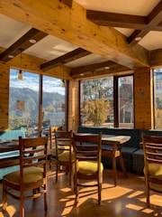 A cozy interior of a wooden cafe with large windows revealing a scenic view of trees and mountains. The room features wooden beams, several wooden chairs with yellow cushions, and tables set for dining. Warm sunlight streams through the windows, casting shadows on the wooden floor.