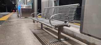 A metal bench is situated on a concrete platform, adjacent to a glass-walled shelter or station. The scene appears to be at night, with artificial lighting and reflections on the glass surfaces. A yellow tactile paving strip runs parallel to the bench, suggesting a public transport setting, likely a train or bus station.