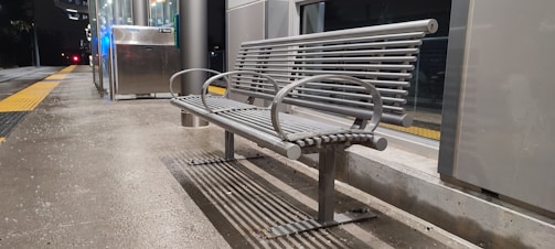 A metal bench is situated on a concrete platform, adjacent to a glass-walled shelter or station. The scene appears to be at night, with artificial lighting and reflections on the glass surfaces. A yellow tactile paving strip runs parallel to the bench, suggesting a public transport setting, likely a train or bus station.