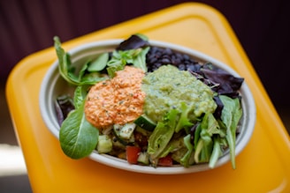 A vibrant salad served in a shallow bowl with a base of fresh greens, topped with dollops of guacamole and a creamy, orange-colored sauce. Dark beans are visible alongside the leafy greens, and there are pieces of cucumber and tomato scattered throughout.