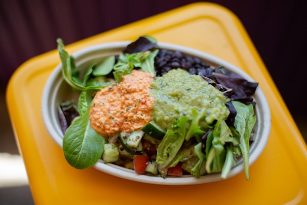 A vibrant salad served in a shallow bowl with a base of fresh greens, topped with dollops of guacamole and a creamy, orange-colored sauce. Dark beans are visible alongside the leafy greens, and there are pieces of cucumber and tomato scattered throughout.