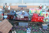 A small toy train set complete with colorful cars is placed outdoors amongst various miniature houses and decorative elements like snowmen. A hedgehog is perched inside a train car labeled 'D & R.G.'. Holiday-themed decorations add a festive atmosphere to the scene.