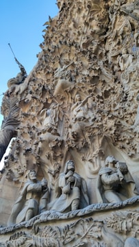 Elaborate stone carvings adorn the facade of a building, featuring intricate depictions of figures in robes holding objects. The stone surface is textured with a natural, organic look, resembling foliage and rock formations.