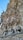 Elaborate stone carvings adorn the facade of a building, featuring intricate depictions of figures in robes holding objects. The stone surface is textured with a natural, organic look, resembling foliage and rock formations.