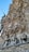 Elaborate stone carvings adorn the facade of a building, featuring intricate depictions of figures in robes holding objects. The stone surface is textured with a natural, organic look, resembling foliage and rock formations.