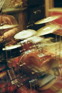 A blurred image captures a drum set with drumsticks in motion, suggesting dynamic movement. The background contains musical accessories and dim lighting, creating a cozy and intimate atmosphere likely in a practice or recording studio.