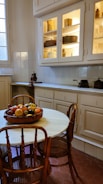 A cozy kitchen with a vintage feel, featuring wooden chairs around a round white table. A fruit bowl filled with a variety of fruits sits on the table. The cabinetry has glass doors with shelves displaying antique-style containers, and the walls are made up of white tiles.