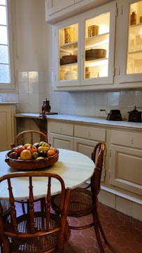 A cozy kitchen with a vintage feel, featuring wooden chairs around a round white table. A fruit bowl filled with a variety of fruits sits on the table. The cabinetry has glass doors with shelves displaying antique-style containers, and the walls are made up of white tiles.
