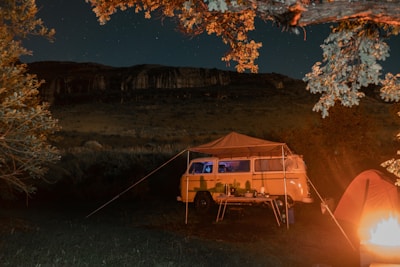 A nighttime camping scene with a classic yellow van set up with an awning and camping table. Warm light from a nearby campfire illuminates the area. A small orange tent is visible, surrounded by trees and a rocky hill in the background under a starry sky.