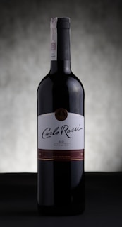 A bottle of Carlo Rossi red wine stands against a softly blurred, neutral background. The label prominently displays the brand name in elegant script, and the bottle is capped with a dark, sleek top.
