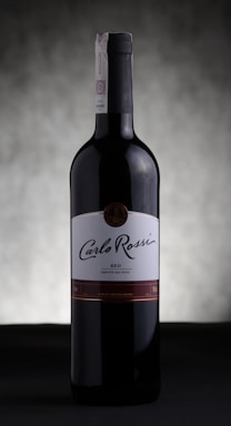 A bottle of Carlo Rossi red wine stands against a softly blurred, neutral background. The label prominently displays the brand name in elegant script, and the bottle is capped with a dark, sleek top.