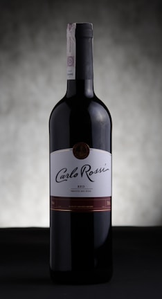 A bottle of Carlo Rossi red wine stands against a softly blurred, neutral background. The label prominently displays the brand name in elegant script, and the bottle is capped with a dark, sleek top.