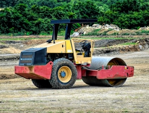 A construction site featuring a heavy-duty road roller machine. The roller is painted yellow and red, with large tires and an open driver's cab. It is positioned on a dirt surface with vegetation and some soil piles visible in the background.
