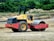 A construction site featuring a heavy-duty road roller machine. The roller is painted yellow and red, with large tires and an open driver's cab. It is positioned on a dirt surface with vegetation and some soil piles visible in the background.