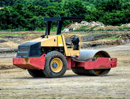 A construction site featuring a heavy-duty road roller machine. The roller is painted yellow and red, with large tires and an open driver's cab. It is positioned on a dirt surface with vegetation and some soil piles visible in the background.