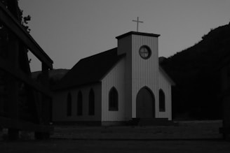 A small, white wooden chapel with a cross on top is set against a backdrop of hills and trees. The building has arched windows and a central circular window.