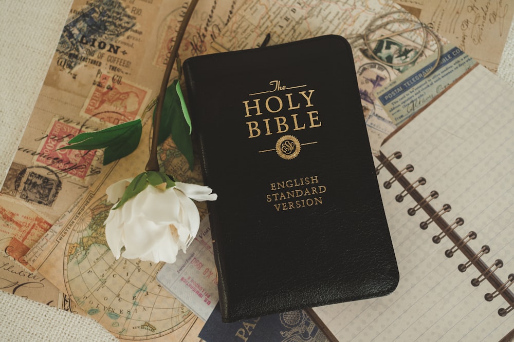 a black bible sitting on top of a table next to a flower