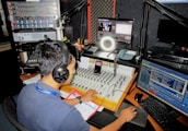 A person wearing headphones operates a sound mixing console in a radio studio. Surrounding them are various pieces of audio equipment, computer monitors displaying different software interfaces, and a microphone setup. There is a calendar and some notes on the desk.