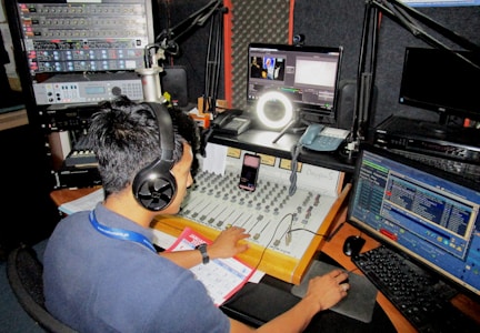 A person wearing headphones operates a sound mixing console in a radio studio. Surrounding them are various pieces of audio equipment, computer monitors displaying different software interfaces, and a microphone setup. There is a calendar and some notes on the desk.