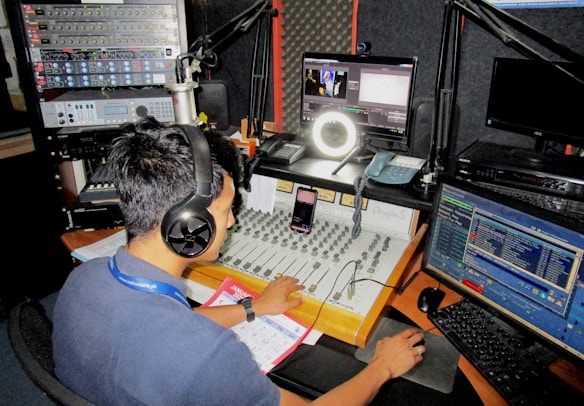 A person wearing headphones operates a sound mixing console in a radio studio. Surrounding them are various pieces of audio equipment, computer monitors displaying different software interfaces, and a microphone setup. There is a calendar and some notes on the desk.
