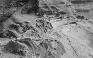 A close-up view of intricate, weathered rock formations displaying smooth, flowing patterns and textures. The geological surface exhibits layers and crevices, highlighting erosion effects.