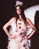 A woman with long brown hair is wearing an ornate feathered white dress with red accents. She dons a silver tiara adorned with feathers, standing with her hands on her hips in front of a spotlight. The lighting creates a dramatic shadow on the wall behind her.