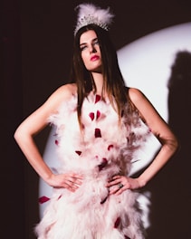 A woman with long brown hair is wearing an ornate feathered white dress with red accents. She dons a silver tiara adorned with feathers, standing with her hands on her hips in front of a spotlight. The lighting creates a dramatic shadow on the wall behind her.