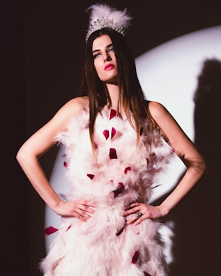 A woman with long brown hair is wearing an ornate feathered white dress with red accents. She dons a silver tiara adorned with feathers, standing with her hands on her hips in front of a spotlight. The lighting creates a dramatic shadow on the wall behind her.