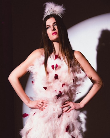 A woman with long brown hair is wearing an ornate feathered white dress with red accents. She dons a silver tiara adorned with feathers, standing with her hands on her hips in front of a spotlight. The lighting creates a dramatic shadow on the wall behind her.