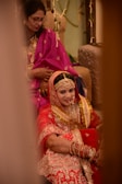 A bride in an ornate red and gold traditional outfit is sitting, adorned with jewelry and henna on her hands. She is accompanied by another woman dressed in a purple sari, who is seated behind her. The setting suggests a wedding ceremony with decorative elements in the background.