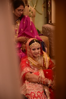 A bride in an ornate red and gold traditional outfit is sitting, adorned with jewelry and henna on her hands. She is accompanied by another woman dressed in a purple sari, who is seated behind her. The setting suggests a wedding ceremony with decorative elements in the background.