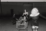 Three women interact playfully with a shopping cart in a dimly lit, outdoor industrial setting. One woman is sitting inside the cart, one is standing on its edge, and another is pulling it, all dressed in fashionable, dark, and edgy outfits.