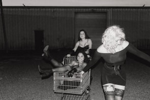 Three women interact playfully with a shopping cart in a dimly lit, outdoor industrial setting. One woman is sitting inside the cart, one is standing on its edge, and another is pulling it, all dressed in fashionable, dark, and edgy outfits.