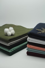 Two neatly stacked piles of folded fabrics in various colors sit on a clean surface. A cotton flower is placed on one pile, with its white fibers contrasting against the green fabric beneath it.