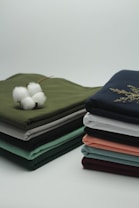 Two neatly stacked piles of folded fabrics in various colors sit on a clean surface. A cotton flower is placed on one pile, with its white fibers contrasting against the green fabric beneath it.