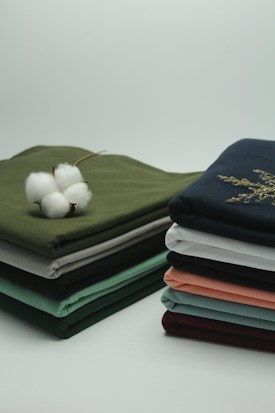 Two neatly stacked piles of folded fabrics in various colors sit on a clean surface. A cotton flower is placed on one pile, with its white fibers contrasting against the green fabric beneath it.