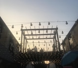 A wooden pergola structure is adorned with numerous hanging light bulbs against a backdrop of a sky transitioning from day to night. People can be seen sitting or standing on the elevated area within the pergola. The scene is softly lit, creating a warm and inviting ambiance.