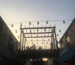 A wooden pergola structure is adorned with numerous hanging light bulbs against a backdrop of a sky transitioning from day to night. People can be seen sitting or standing on the elevated area within the pergola. The scene is softly lit, creating a warm and inviting ambiance.