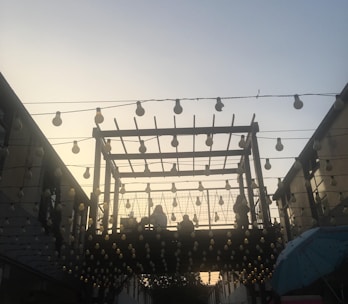 A wooden pergola structure is adorned with numerous hanging light bulbs against a backdrop of a sky transitioning from day to night. People can be seen sitting or standing on the elevated area within the pergola. The scene is softly lit, creating a warm and inviting ambiance.