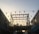 A wooden pergola structure is adorned with numerous hanging light bulbs against a backdrop of a sky transitioning from day to night. People can be seen sitting or standing on the elevated area within the pergola. The scene is softly lit, creating a warm and inviting ambiance.
