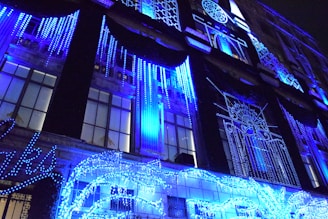 A building facade is adorned with vibrant blue holiday lights, forming intricate patterns and designs. The lights drape down in vertical strands, creating a mesmerizing visual display. The structure features large windows that reflect the glowing decorations, enhancing the festive atmosphere.