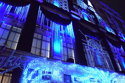 A building facade is adorned with vibrant blue holiday lights, forming intricate patterns and designs. The lights drape down in vertical strands, creating a mesmerizing visual display. The structure features large windows that reflect the glowing decorations, enhancing the festive atmosphere.