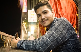 A young man in a checkered shirt is leaning on a table while holding a smartphone. Behind him, there's a bright sign, some glowing lights, and red drapery. The setting appears to be outdoors in a lively or urban environment, possibly near a restaurant or shop entrance.