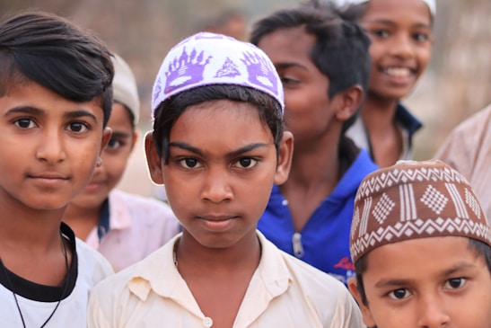 A group of boys, some wearing traditional caps, standing closely together with a blurred background. Their expressions range from serious to cheerful.